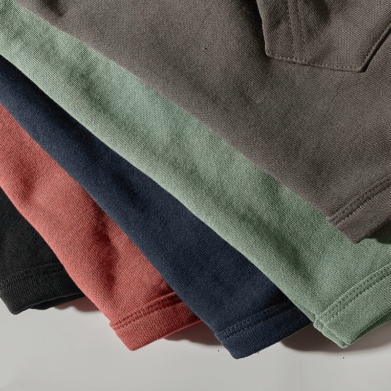 Stack of solid color sweatshirts with kangaroo pockets in brown, green, red, and blue hues, perfect for warm, casual wear.